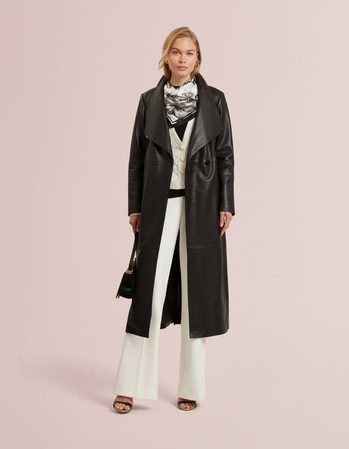 Woman wearing a black coat, white pants, and a patterned scarf on a light gray background